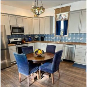 Modern kitchen remodel with white cabinetry, blue tile backsplash, stainless steel appliances, and a wooden table with plush chairs.